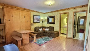 Una sala de estar acogedora con piso de madera, un sofá, una mesa de centro y una mesa de comedor. Se ve una cocina a través de una puerta abierta.