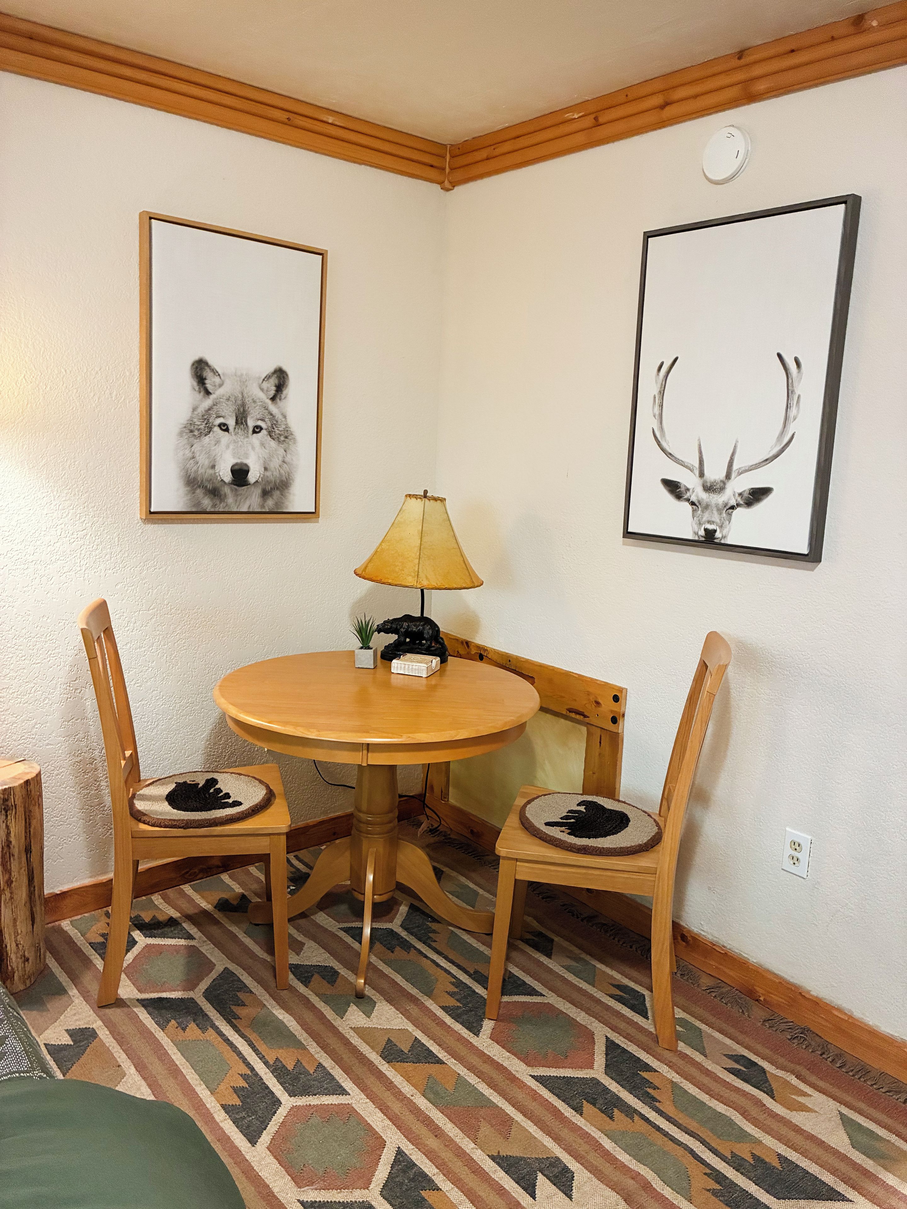 Un comedor con una mesa redonda de madera, dos sillas, una lámpara y dos cuadros en la pared.