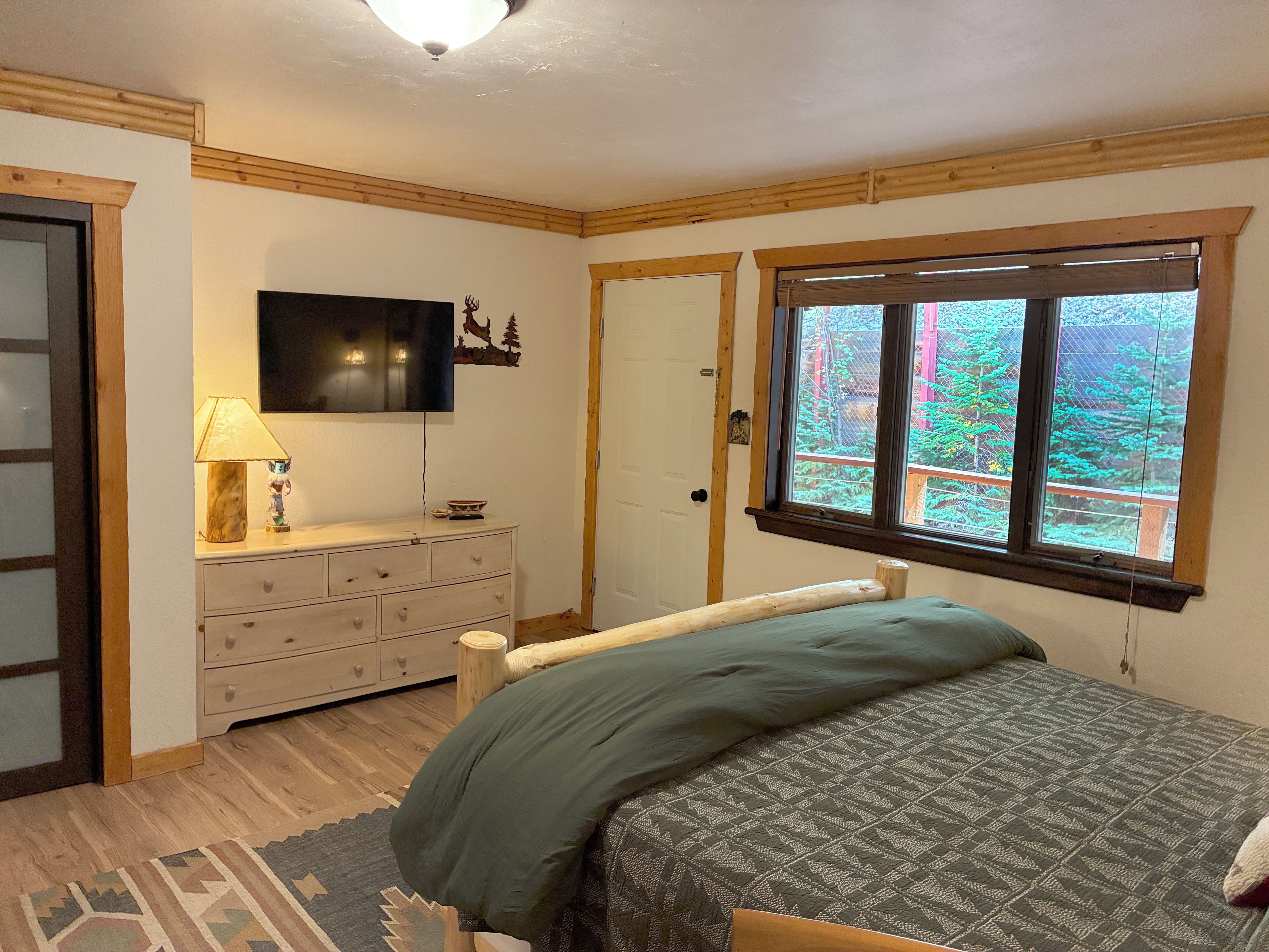 A bedroom with a bed, dresser, television, and a window with a view of trees.