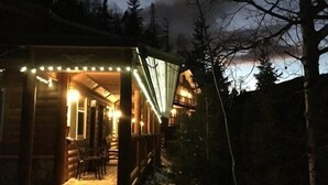 En træhytte med en overdækket veranda og lyskæder, omgivet af træer og sne.