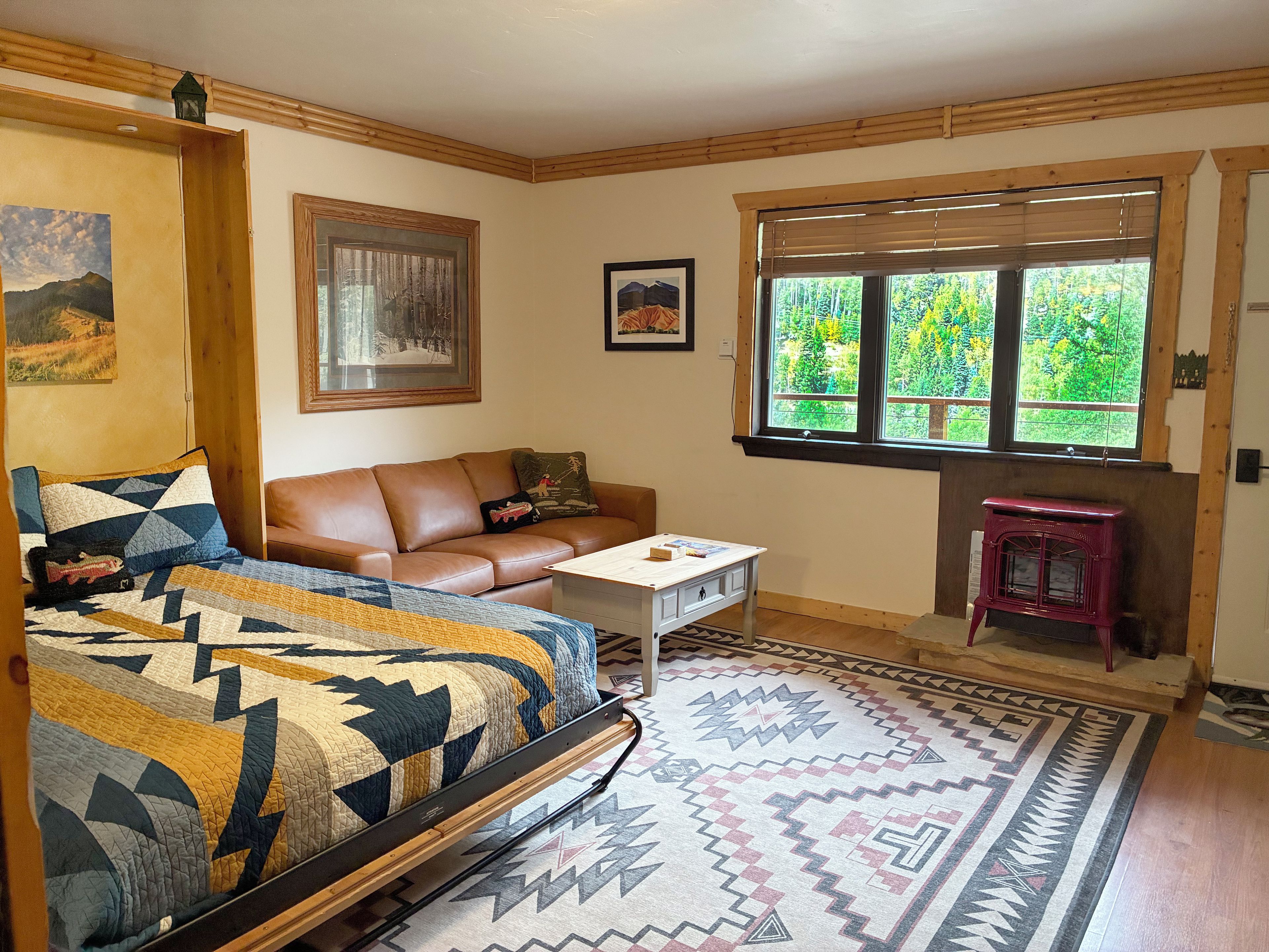 A cozy room with a bed, a brown leather sofa, a small table, and a window with a view of greenery.