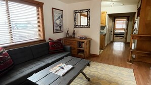 Ein Wohnzimmer mit einem dunklen Sofa, einem Holztisch und einem gemusterten Teppich. Im Hintergrund ist eine Küche mit Holzschränken und einem Fenster mit Jalousien zu sehen.