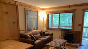 A cozy living room with a brown leather couch, a wooden coffee table, and a striped bench. A fireplace is visible in the background, and there is a large window with blinds.