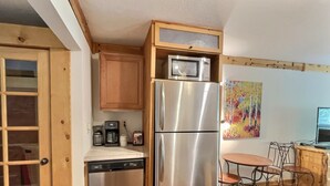 Eine kleine Küche mit Edelstahlkühlschrank, einer Mikrowelle und einem Geschirrspüler. Eine Holztür mit Glaseinsätzen, ein kleiner Tisch mit Stühlen und ein farbenfrohes Bild an der Wand.