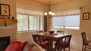 A cozy dining area with a wooden table, chairs, and a chandelier. There is a fireplace on the left and a window with curtains. - High Altitude Gem at Taos Ski Valley (Taos Ski Valley)