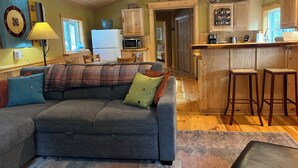 A cozy living room with a gray sofa, wooden ceiling, and a kitchen area with wooden cabinets and a bar stool. - Cozy Amizette - Cabin (Taos Ski Valley)