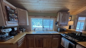 A wooden kitchen with a built-in stove, a coffee maker, and a window with a view of trees. - Cozy Amizette - Cabin (Taos Ski Valley)
