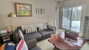 Ein gemütliches Wohnzimmer mit grauem Sofa, einem Holztisch und einem großen Fenster mit Blick auf Schnee.