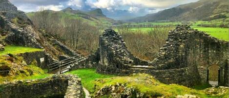 Ancient stone ruins with a pathway leading through them, surrounded by green fields and mountains.