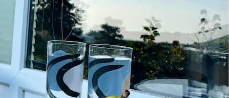 A bowl of fruit and two glasses on a table by a window.
