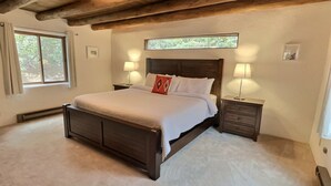 A bedroom with a large bed, wooden headboard, two bedside tables with lamps, a window with curtains, and a wooden ceiling. - Beautiful Adobe in the Pines Above Taos (Taos)