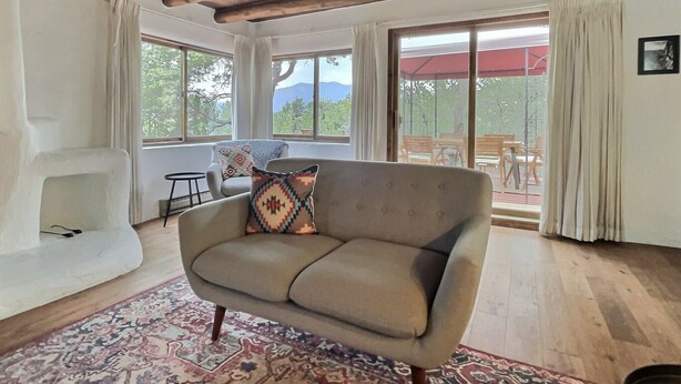 A cozy living room with a beige sofa, patterned cushions, a wooden ceiling, large windows with a view of the outdoors, and a patterned rug. - Beautiful Adobe in the Pines Above Taos (Taos)