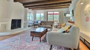 A spacious living room with a fireplace, large windows, and a high wooden ceiling. - Beautiful Adobe in the Pines Above Taos (Taos)