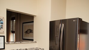 A kitchen with a stainless steel refrigerator, dark cabinets, a marble countertop, and a wooden floor.