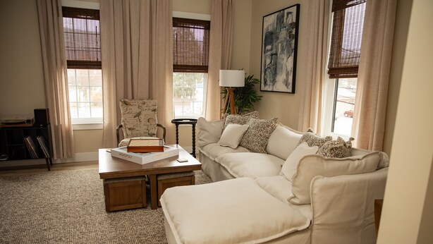 A living room with a beige sofa, a coffee table, and a chair. There is a bookshelf with books and a lamp.