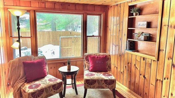A cozy room with wooden walls and a large window. There are two upholstered chairs with floral patterns and a small round table. A lamp is on the left side, and a shelf with a 'Thankful' sign is on the right. - Cozy 3 b/r home w/ quick access to snowmobile trails in the beautiful Northwoods (Minocqua)