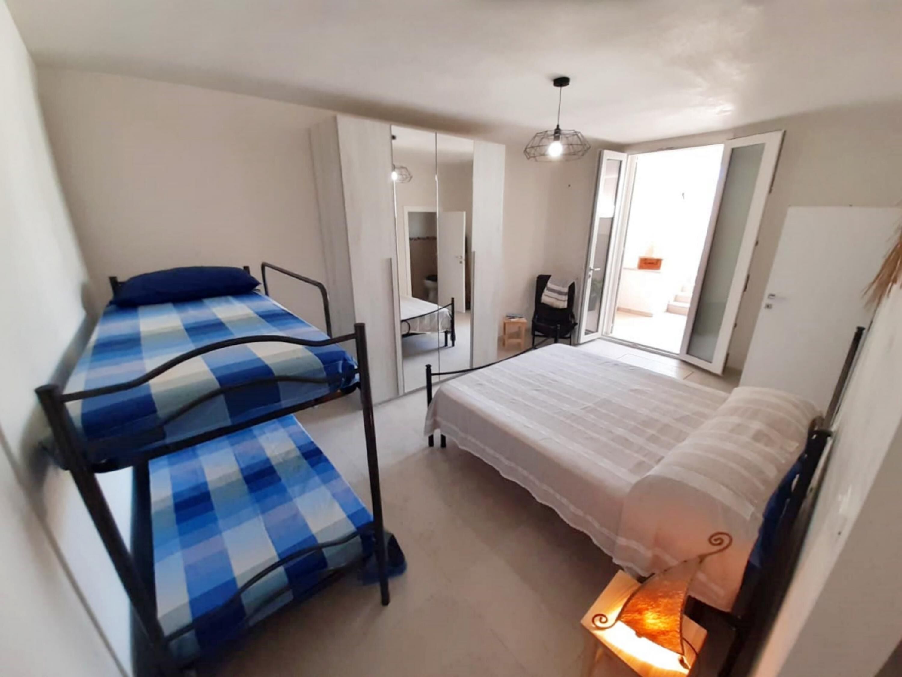Habitación tipo dormitorio con dos literas, un espejo grande, una lámpara en el techo y una puerta corrediza de vidrio que da a un balcón.