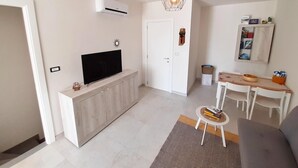 A modern living room with a flat-screen TV on a wooden cabinet, a dining table with chairs, a patterned rug, and a pendant light.