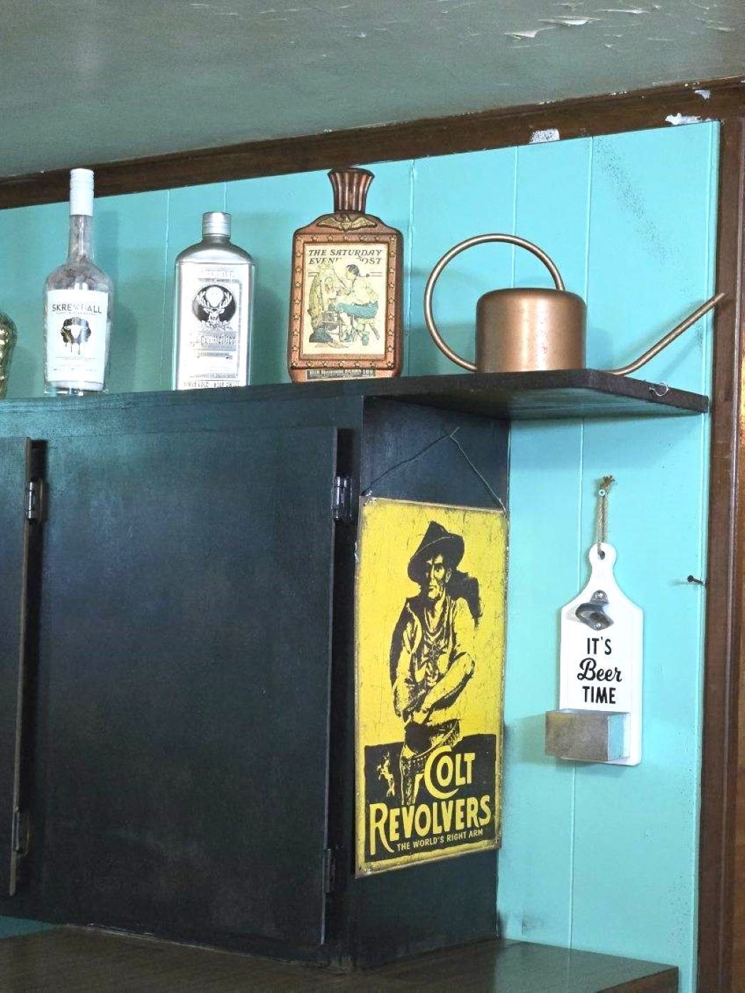 Una cocina con un armario negro, una repisa con botellas, un cartel vintage y una olla de metal.