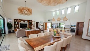 A spacious open-plan living area with a large wooden dining table surrounded by chairs, a kitchen area with modern appliances, and a large TV on a wooden cabinet.