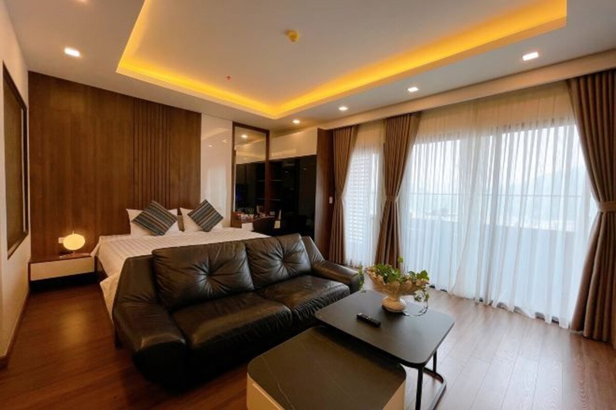 A modern hotel room with a bed, a leather sofa, a small table, and a large window with curtains.