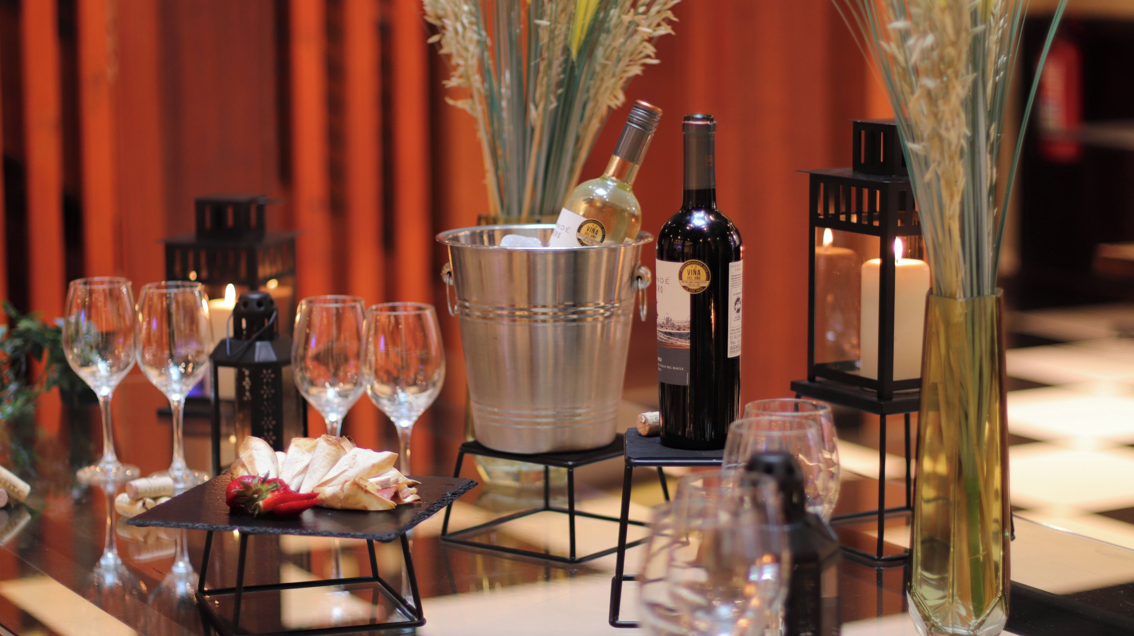 A table set with wine glasses, a bottle of wine, a bucket with a bottle, and a plate with food.