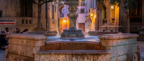 A historic fountain with a central basin and four surrounding steps, surrounded by old buildings with balconies and warm lighting.
