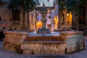 A historic fountain with a central basin and four surrounding steps, surrounded by old buildings with balconies and warm lighting.