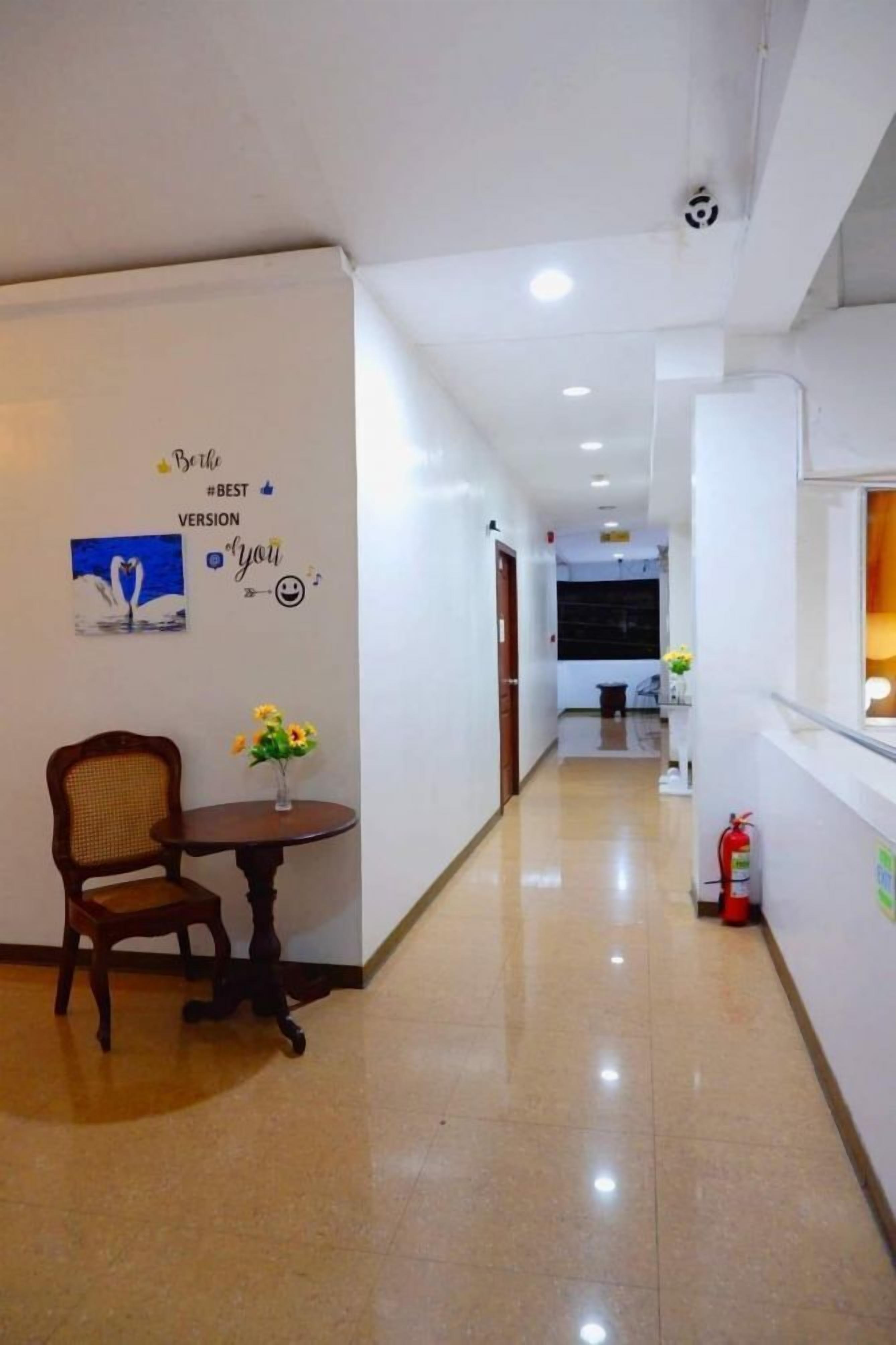 Un pasillo de hotel con una silla de madera y una mesita con flores, un extintor y una pared con texto decorativo y una imagen con forma de corazón.