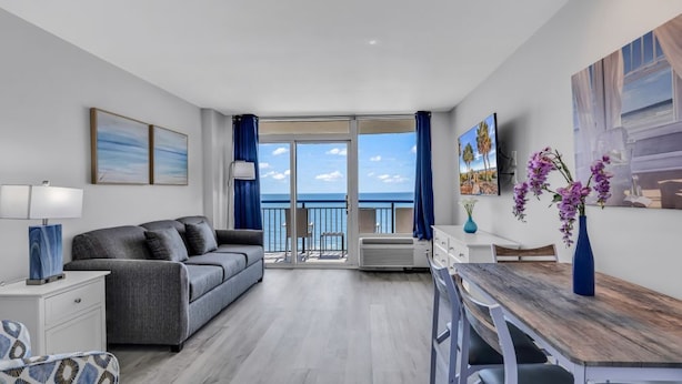 A modern room with a grey sofa, a wooden dining table, and a balcony with a view of the sea. - Boardwalk Resort: Ocean Breeze Retreat (Myrtle Beach)