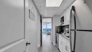 A narrow hallway with a kitchen area on the right, featuring a refrigerator, microwave, and oven. A door on the left leads to another room. - Boardwalk Resort: Ocean Breeze Retreat (Myrtle Beach)