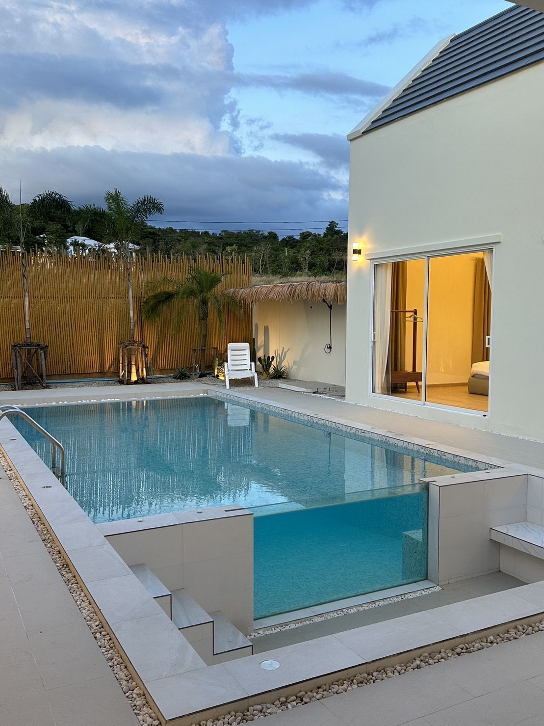Una piscina rettangolare con bordo piastrellato, circondata da una recinzione in bambù e palme.