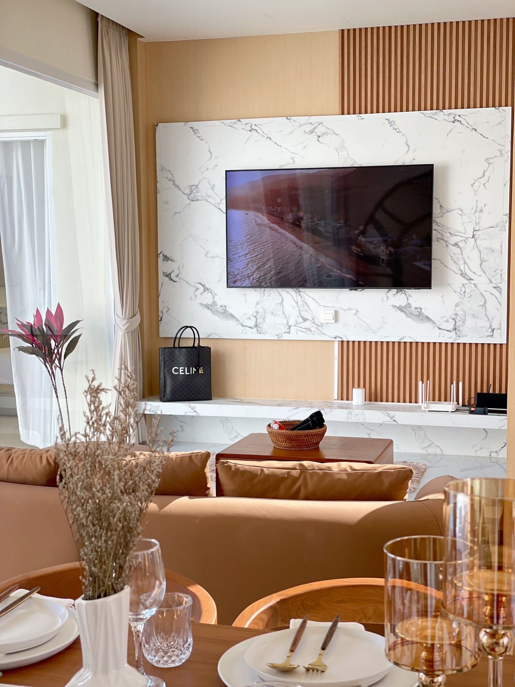 A modern living room with a large flat-screen TV mounted on a marble wall, a sofa, a dining table set with plates and glasses, and a decorative vase with dried flowers.