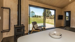 Un interior moderno con un ventanal grande, una estufa de leña empotrada y una zona de comedor con un bol en la mesa.