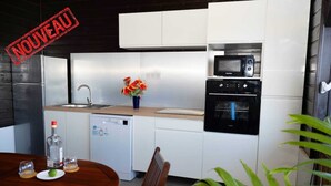 A modern kitchen with white cabinets, a built-in microwave, and an oven. There is a sink, a dishwashing machine, and a dining table with chairs. A bottle and glasses are on the table, and a vase with flowers is on the counter. - F3 Hibiscus - vue mer - Piscine Chauffée - 9 min de la mer (La Trinité)