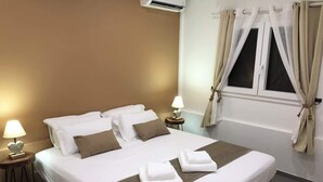 A neatly made bed with white and brown bedding, two bedside lamps, a window with curtains, and an air conditioning unit above the bed. - F3 Hibiscus - vue mer - Piscine Chauffée - 9 min de la mer (La Trinité)