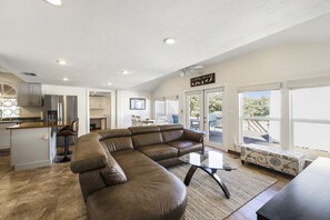 A modern living room with a brown leather sectional sofa, a glass coffee table, and a kitchen area with stainless steel appliances.