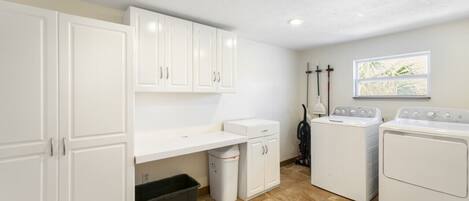 A laundry room with white cabinets, a washing machine, a dryer, and a trash bin.