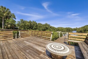 A wooden deck with benches and a circular table, overlooking a body of water and surrounded by greenery.