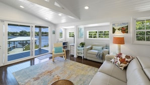 A spacious living area with a large sliding glass door leading to a deck overlooking a body of water. There is a white sofa, a chair, and a table with a lamp.