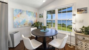 A dining area with a round table, white chairs, a large abstract painting, and a view of the ocean through a large window.