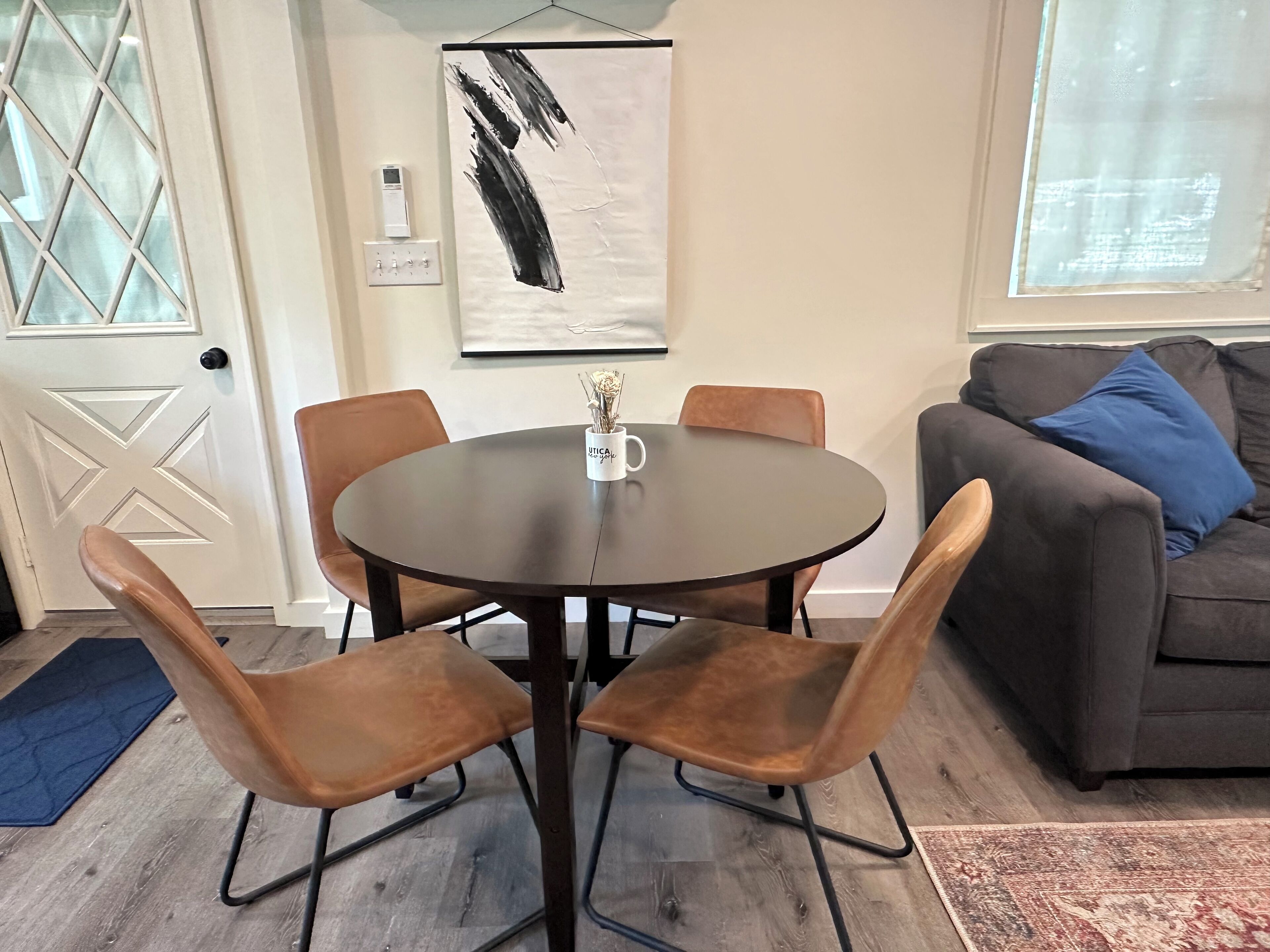 Un coin repas avec une table ronde, quatre chaises et une tasse à café. Il y a un tableau au mur, un thermostat et une fenêtre avec des rideaux.