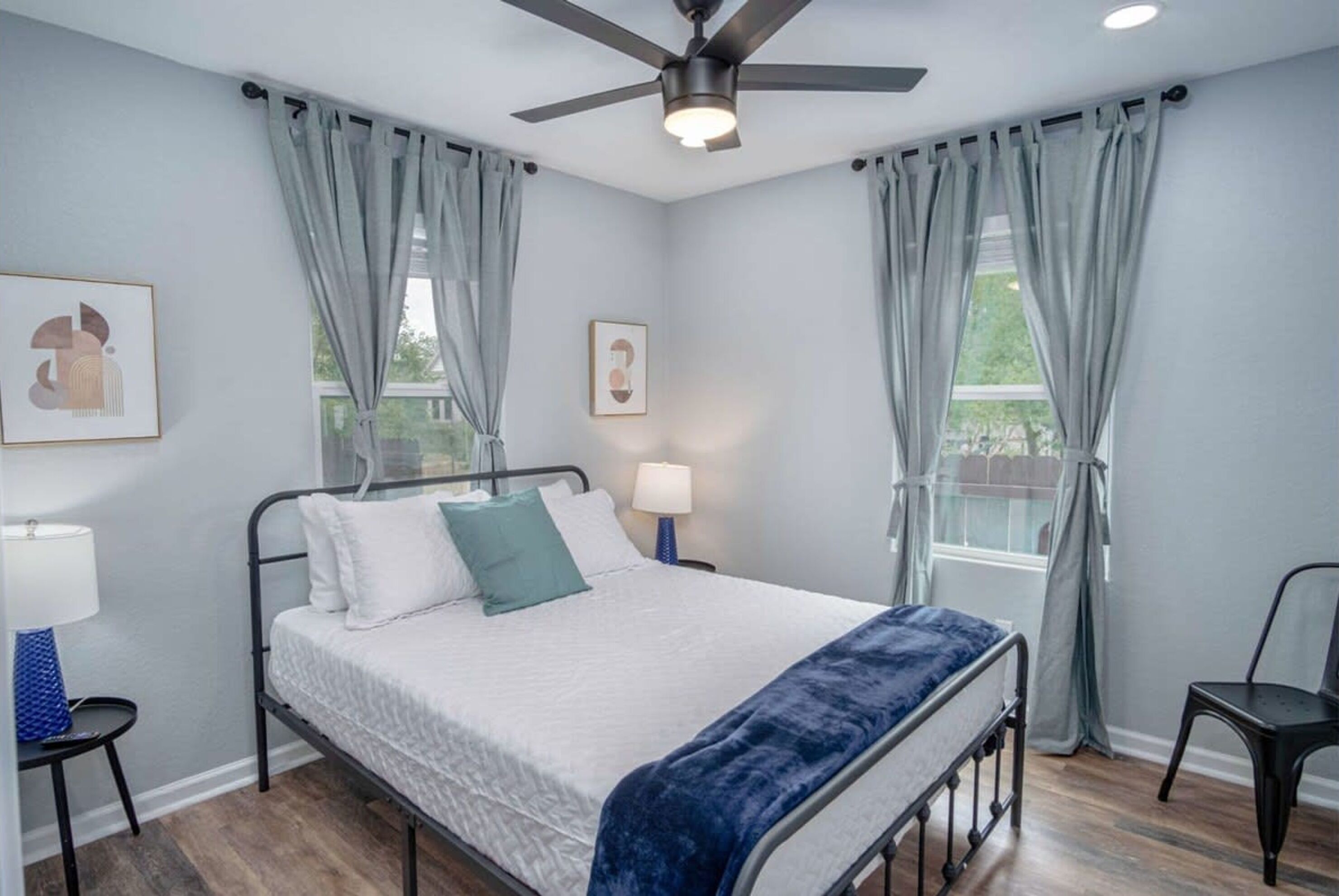 A bedroom with a bed, two bedside lamps, a ceiling fan, and two windows with curtains.
