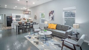 A modern living room with a gray sofa, a coffee table, and a dining area with a kitchen in the background. - Captivating 4BR 2BA Home Near Awesome Downtown (San Antonio)