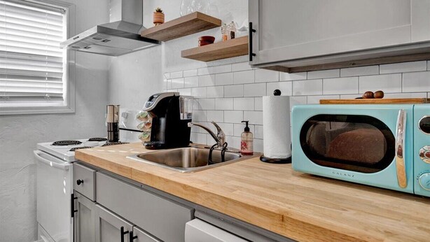A modern kitchen with white cabinets, a wooden countertop, a stainless steel sink, a coffee maker, a microwave, and a window with blinds. - OSU Weekend Retreat - Steps to Campus (Stillwater)