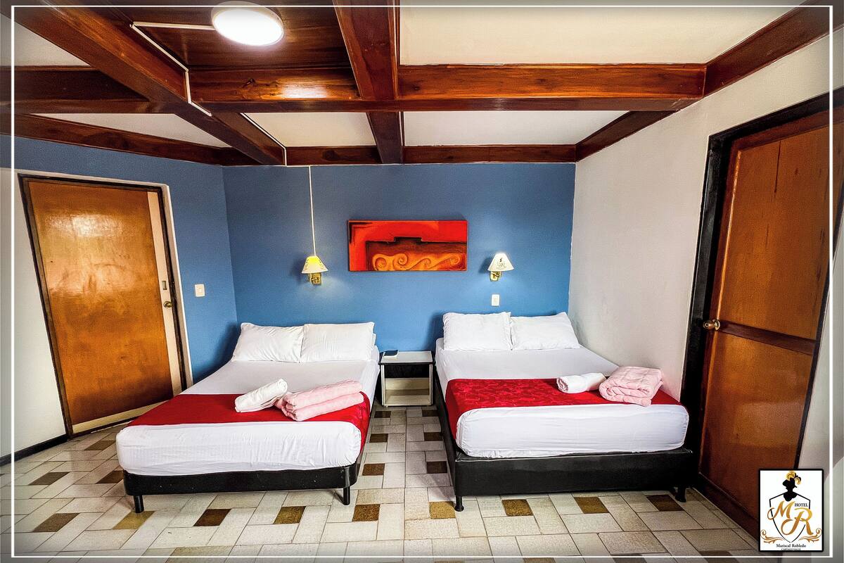 A hotel room with two beds, wooden ceiling beams, a blue wall, and a red decorative piece above the beds.