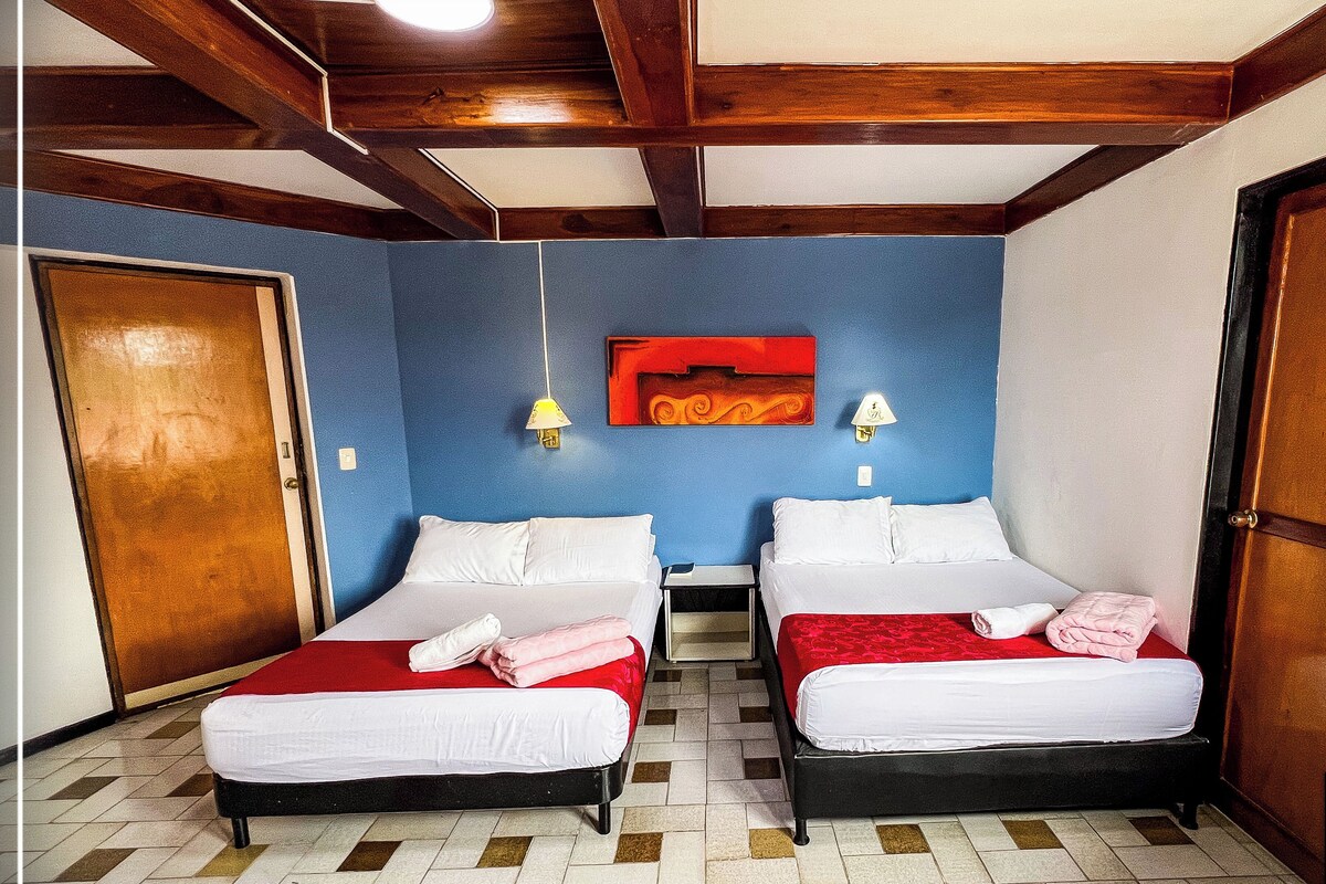 Camera d'albergo con due letti, travi in legno al soffitto, una parete blu e un elemento decorativo rosso sopra i letti.