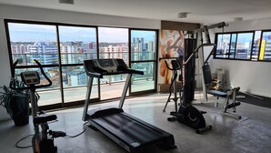A gym with cardio equipment, weight machines, and a large window with a city view. - Grande Apartamento Beira-Mar (Maceió)