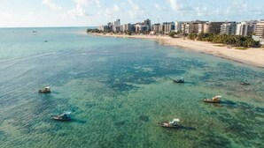A coastal cityscape with clear turquoise waters, several boats, and a beachfront hotel area. - Grande Apartamento Beira-Mar (Maceió)
