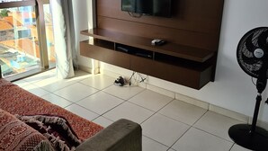 A modern living room with a flat-screen TV mounted on a wooden wall, a sofa with a patterned throw, a standing fan, and a view of a cityscape through a large window. - Grande Apartamento Beira-Mar (Maceió)